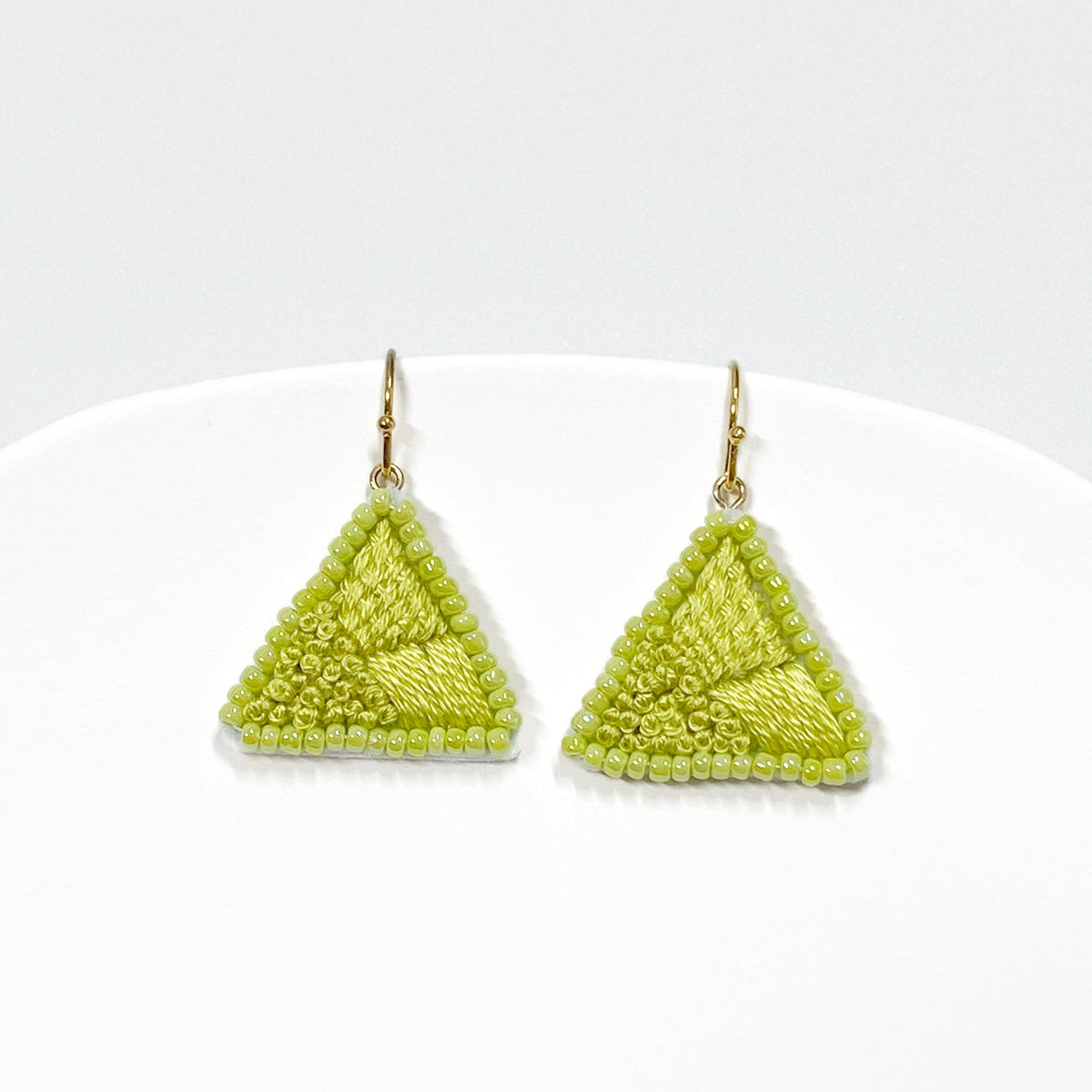 A pair of handmade triangular beaded earrings in a vibrant lime green color, suspended from gold-toned hooks, displayed on a white curved stand against a light gray background.