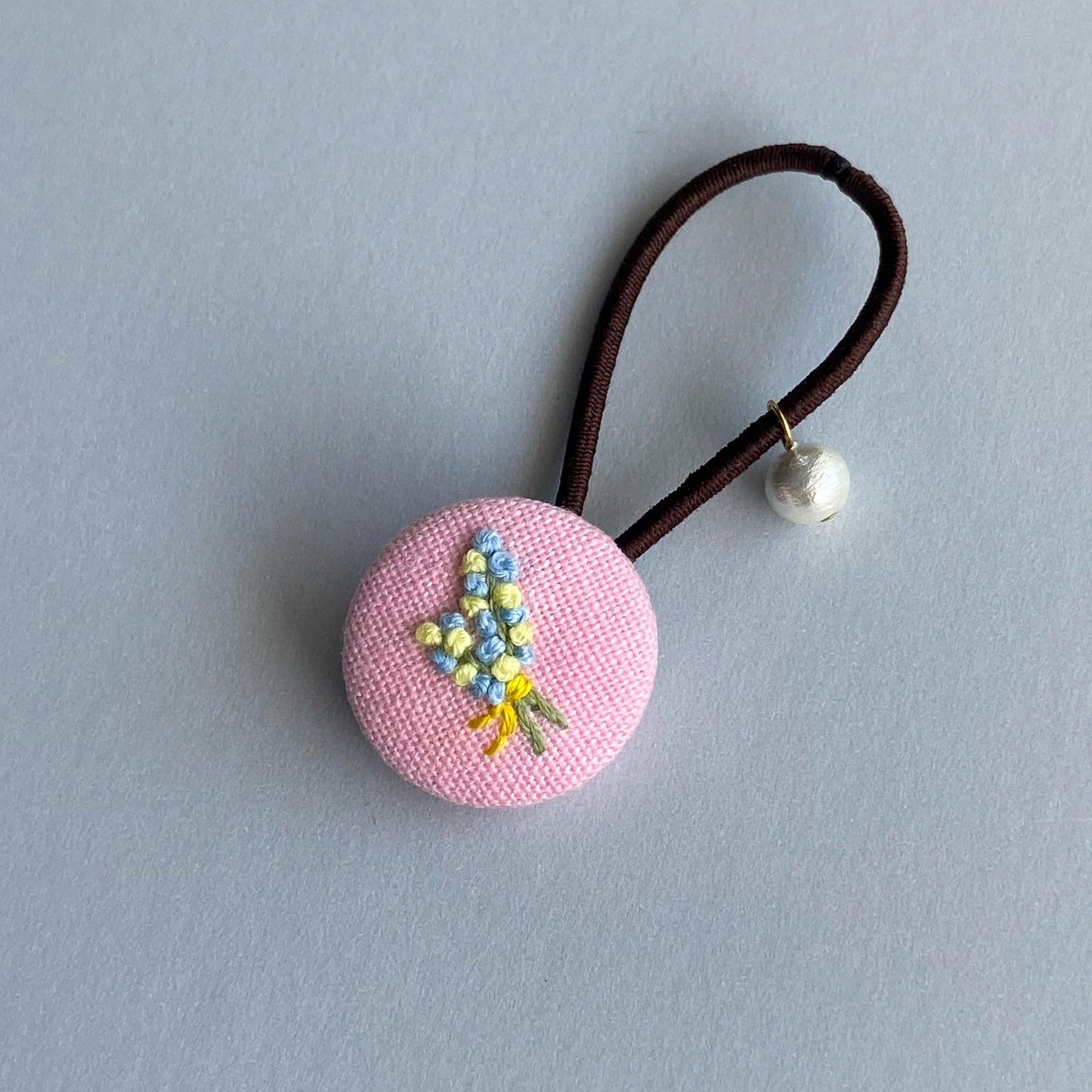 A pink fabric-covered button hair tie with a small embroidered floral design and a dangling pearl bead.