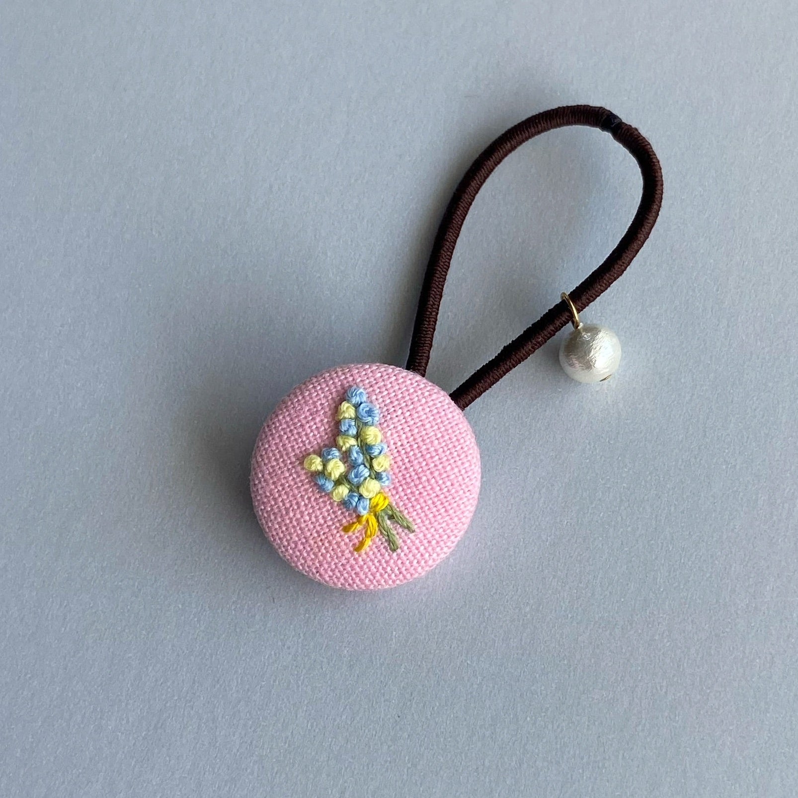 A pink fabric-covered button hair tie with a small embroidered floral design and a dangling pearl bead.