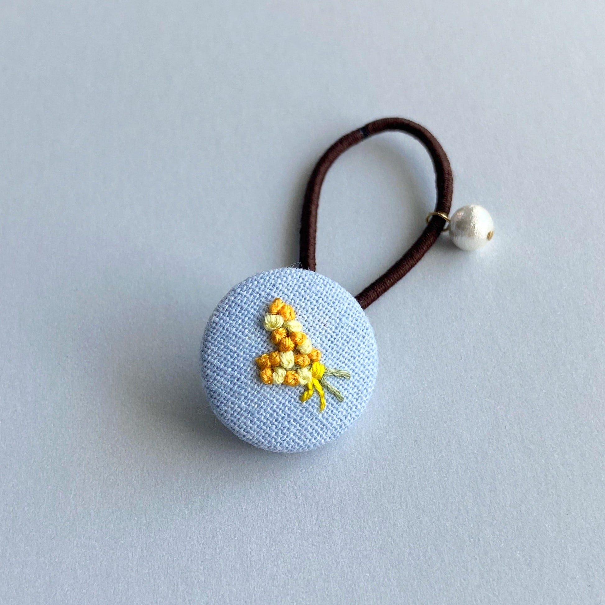 A blue fabric-covered button hair tie with a small embroidered floral design and a dangling pearl bead.