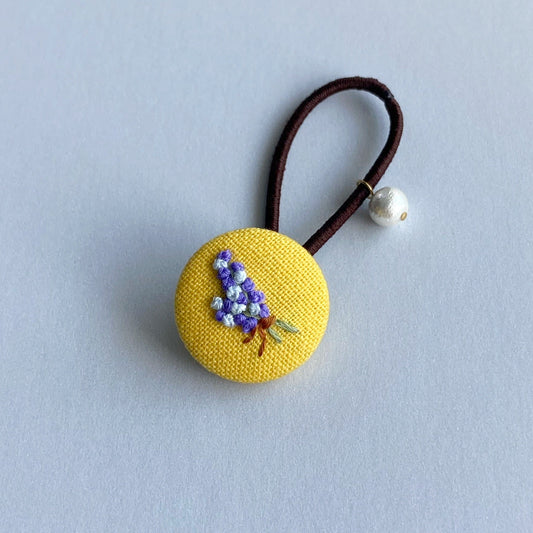 A pink fabric-covered button hair tie with a small embroidered floral design and a dangling pearl bead.
