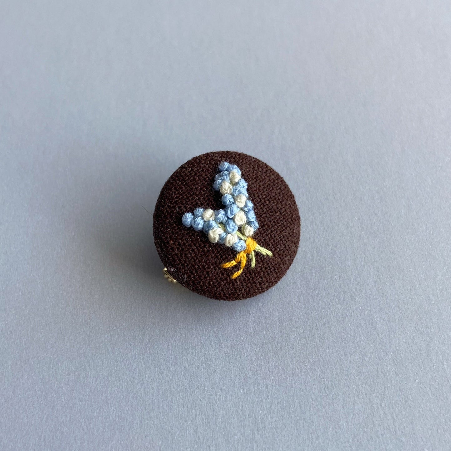 A round brown fabric pin with a small embroidered floral design.