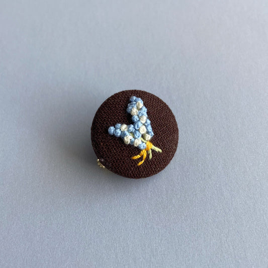 A round brown fabric pin with a small embroidered floral design.