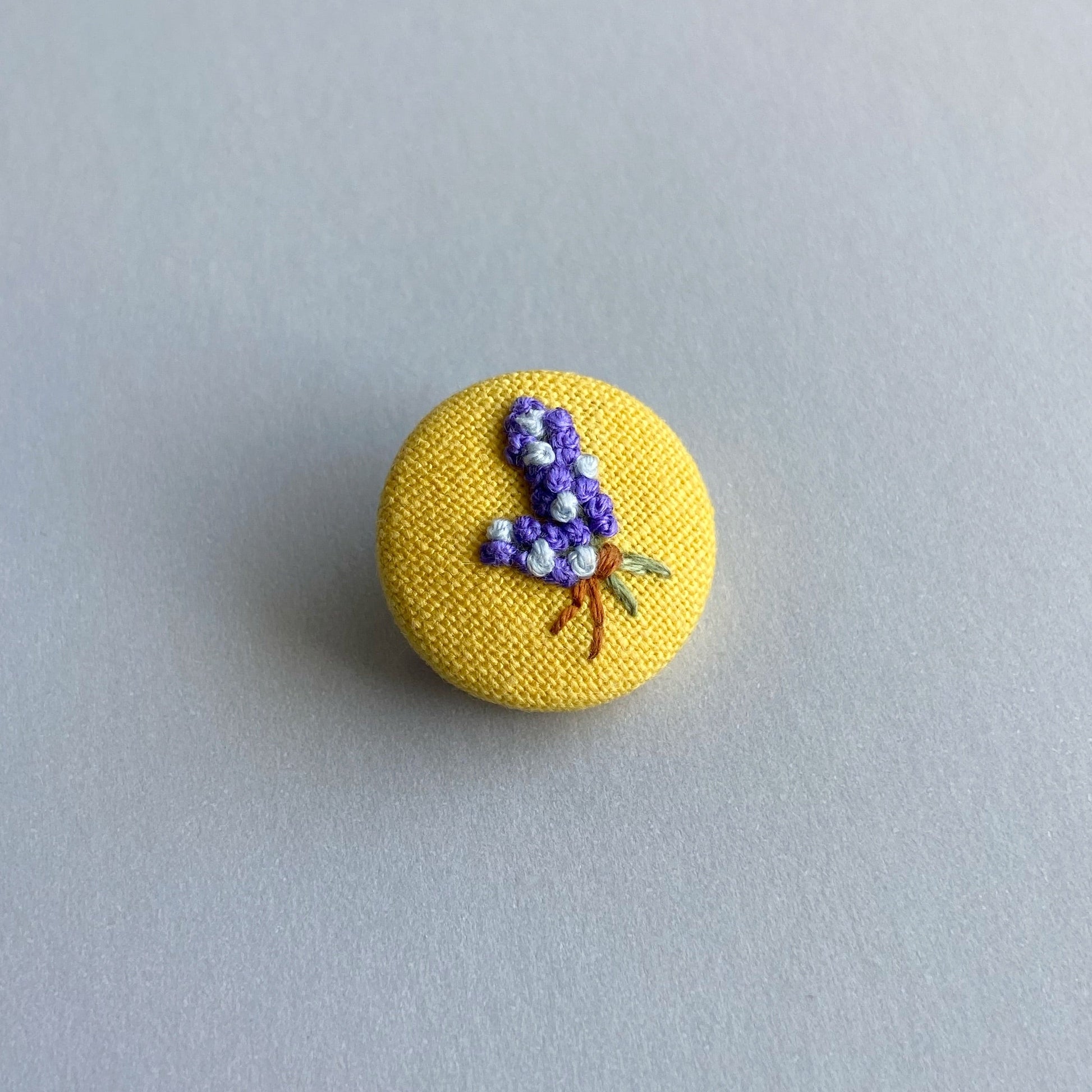 A round yellow fabric pin with a small embroidered floral design.
